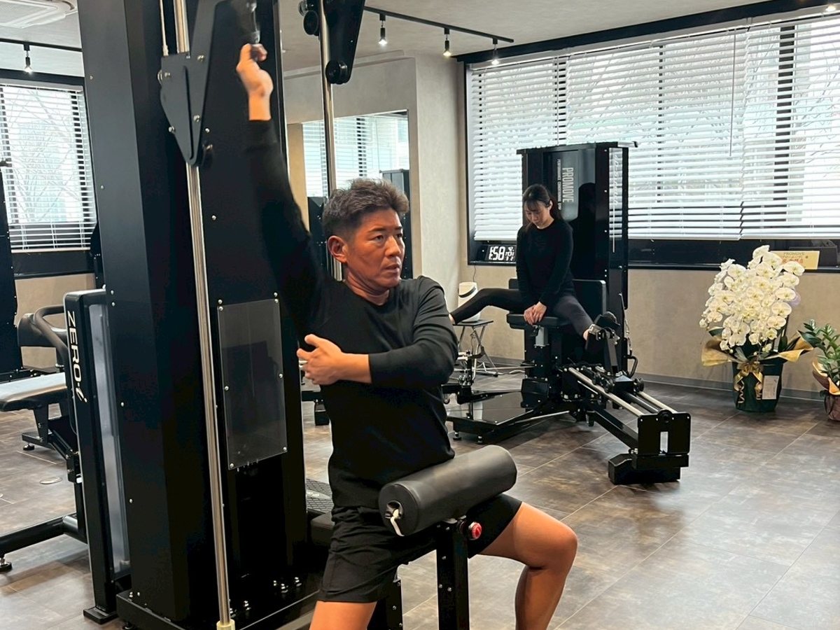40〜50 代の男性会員様がトータルショルダージョイントで肩まわりを動かしている様子。奥では別の会員様がマシンを使っている